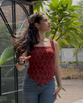 A woman wearing a red sleeveless top with a black floral pattern and blue jeans, standing outdoors next to a glass-walled structure with green plants in the background.