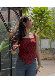 A woman wearing a red sleeveless top with a black floral pattern and blue jeans, standing outdoors next to a glass-walled structure with green plants in the background.