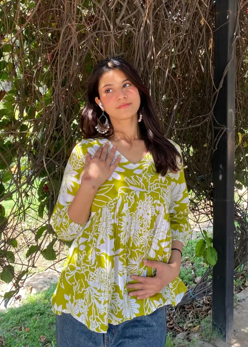 Woman in a yellow floral top standing in front of a natural background with trees and plants.